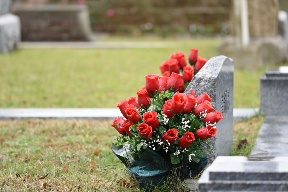 A bouquet of fresh red roses arranged with small white filler flowers and green foliage, wrapped in dark green cellophane, resting against a grey granite headstone in a cemetery. The roses are in full