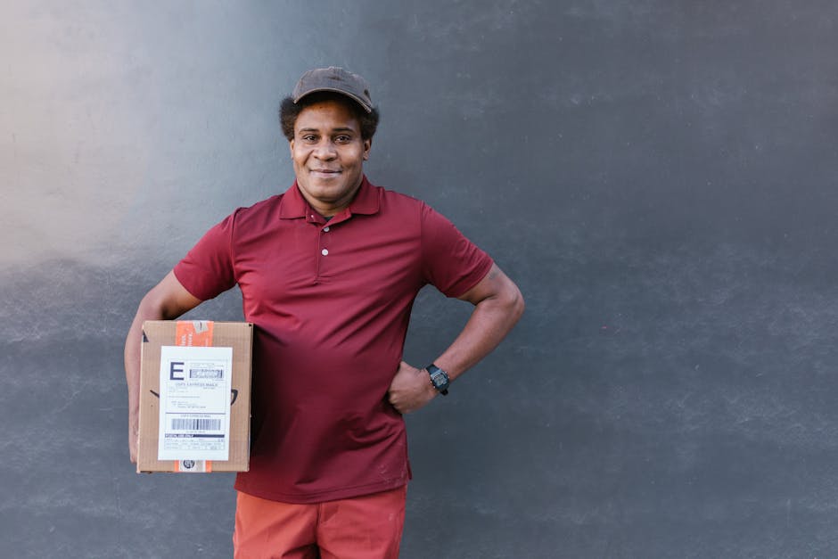 A friendly male delivery person with dark curly hair, wearing a dark grey cap and a maroon polo shirt, stands outdoors against a dark grey wall. He is holding a brown cardboard parcel with a shipping