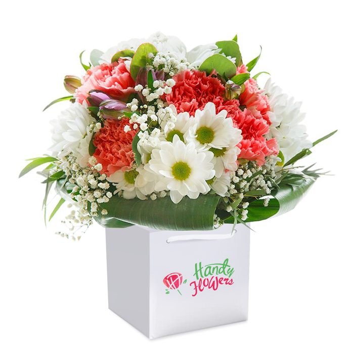Set against a spotless white backdrop, this vibrantly arranged bouquet is a celebration of colour and texture that calls to mind the spirited character of Canning Town. The focal blooms-lush, coral-hued carnations-display exquisitely frilled petals, each shade softly melting into the next, bringing warmth and movement to the composition. Scattered among them, daisy-like white chrysanthemums with fresh, lime-green centres shine with cheerful clarity, reminiscent of spring mornings at Trinity Gardens. The dainty, lightweight sprigs of baby's breath weave their fine white blossoms through the bouquet, creating a cloud-like softness that gently cushions and elevates the primary flowers. Nestled discreetly within the arrangement are several closed buds, lightly flushed with violet and pink, suggesting anticipation and the ongoing beauty of renewal-perfect for new beginnings or celebrations. The bouquet's structure is defined by an inviting array of glossy, dark green foliage and broad leaves that arc around the blooms, creating balance and an impression of abundance. It's thoughtfully presented in a square white gift bag, practical with crisp cord handles and ready for same-day surprises across Canning Town or a delivery near Rathbone Market. Every flower is expertly positioned for maximum visual harmony and freshness, representing the skilled touch of community florists committed to quality. This image embodies the joy and sincerity of gifting blooms locally, capturing both the artistry of the craft and the genuine care of Florist Canning Town.