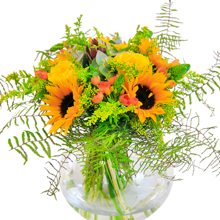 This exuberant bouquet, inspired by the energy of Canning Town and its lively markets, features a captivating mix of golden sunflowers at centre stage. Their broad, cheerful faces stand proudly above swirls of smaller yellow filler flowers-perhaps mimosa or solidago-echoing the vibrancy found near Rathbone Market. Between these sunny elements, hints of soft coral-orange blooms emerge, their cup-like shapes whispering a soft warmth evocative of a sunset over the Thames Barrier Park. Glossy green stems and abundant foliage form the framework of this arrangement; feathery fern fronds stretch gracefully outward, interlacing with modern, silvery eucalyptus leaves and plump lime-green greens for depth and movement. All are gathered in a pristine spherical glass vase, the water within catching the daylight and making every colour sing. Whether destined as a bright centrepiece on a Silvertown family table or a comforting gesture for a loved one nearby, the bouquet's rich textures and sweet hints of earthy perfume kindle a sense of joy and renewal. Thoughtfully created by Canning Town's experienced floral artisans, this arrangement is perfect for moments of celebration, encouragement, or simple acts of kindness, offering a splash of local sunshine and heartfelt craftsmanship to any recipient.