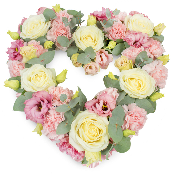 A generously proportioned heart-shaped wreath, designed as a funeral or memorial tribute, appears against a spotless white background, every detail of the floristry clearly visible. The heart is completely filled with flowers, creating a plush, continuous surface where the individual blooms blend together yet keep their own character. Cream and pale yellow roses are placed thoughtfully around the shape, their tightly furled centres swirling out into velvety, full petals that catch the light and suggest a sense of calm dignity. Between these roses lie clusters of delicate pastel pink carnations, their frilled and layered petals adding a soft, almost feathered texture that lends the arrangement a gentle, comforting presence. Here and there, deeper pink and mauve lisianthus blossoms open more loosely, their slightly cupped, ruffled petals introducing a richer tone and a graceful, almost poetic movement through the design. Interlaced among the flowers, slender stems of eucalyptus with rounded, silvery-green leaves create a soothing backdrop, their cool colour palette balancing the warmth of the creams and pinks. Small yellow-green buds emerge subtly from the foliage, hinting at fresh growth and continuity. The composition feels lush and abundant, with a carefully balanced symmetry that makes the heart shape clearly defined yet soft-edged. It evokes the quiet atmosphere of a farewell service in Canning Town, perhaps at a local chapel or crematorium, where flowers offer comfort when words feel inadequate. This heart-shaped wreath would sit beautifully beside a casket or at the front of a room, expressing sympathy, remembrance, and enduring affection for someone deeply loved.