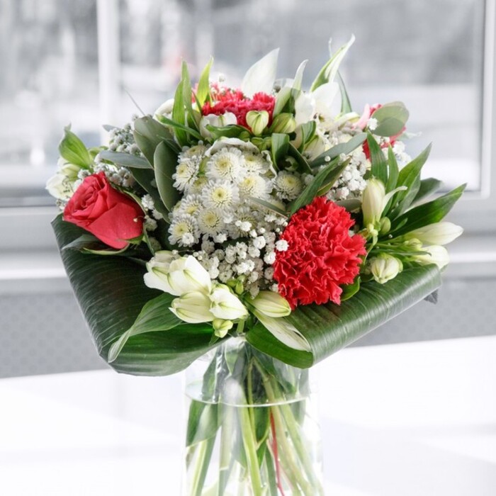 A luxurious red and white bouquet commands attention from the centre of the frame, carefully arranged in a clear cylindrical glass vase that showcases the straight, freshly cut green stems below the waterline, like a miniature garden rooted in crystal. The composition is rich with romantic detail: deep red roses, velvety and full, spiral open alongside a generously ruffled crimson carnation that forms a prominent focal point at the front. Around these passionate tones are gentle white counterpoints-elegant white alstroemeria with slim, speckled petals, rounded white chrysanthemum-type blooms, and several plump white lily buds still tightly closed, promising a slow unfurling over the coming days. Tiny clusters of white gypsophila drift between the larger flowers, creating an airy, cloud-like halo that softens the stronger shapes and adds a sense of celebratory sparkle. The base of the bouquet is wrapped in broad, glossy, dark green leaves, curled and layered into an elegant collar that cradles the blooms and gives the arrangement a clean, contemporary outline, perfect for a stylish living room near Star Lane or a restaurant table close to the Royal Docks. Taller eucalyptus-style sprigs and pointed green foliage rise through the flowers, adding height, movement, and subtle variations of green that evoke a fresh walk around nearby parks and riverside paths. The bouquet rests on a smooth white surface against a softly blurred, light-filled background, suggesting daylight pouring in through a window in a Canning Town home or office. The lighting is gentle yet bright, highlighting textures and colours without harsh shadows. This arrangement feels both modern and timeless-an ideal centerpiece for anniversaries, engagement dinners, or heartfelt congratulations delivered across the E16 area.