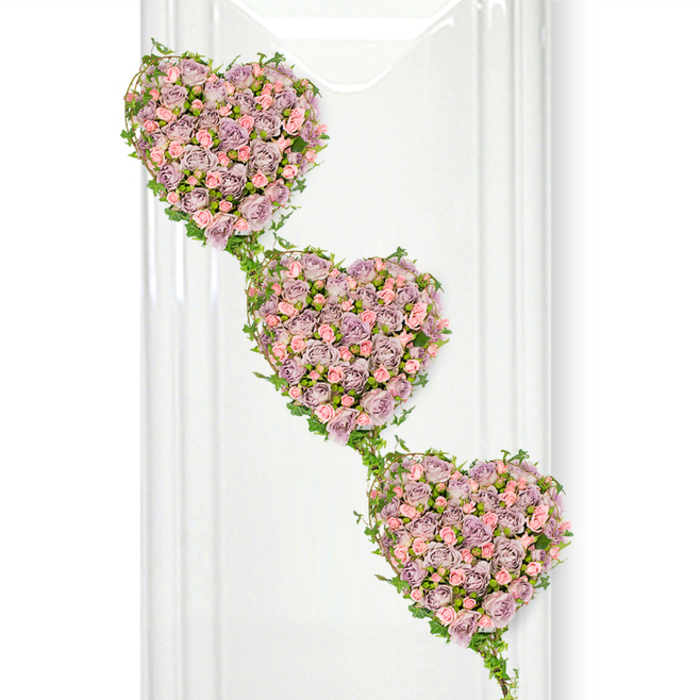 This floral image shows a beautifully crafted sympathy tribute of three cascading heart shapes, designed with the care and precision you would expect from an experienced Canning Town florist. Each heart is fully outlined and filled with closely set roses in soothing funeral tones of dusty lavender, soft mauve, and peppermint-tinged pastel pink. The roses are mainly small to medium in size, arranged so tightly that they create an almost quilted effect, every petal overlapping the next like layers of cherished memories. Subtle variations of purple and pink ripple through the hearts, giving depth and texture, with a mix of open blooms and half-open buds suggesting continuity and the passage of time. Fresh green ivy twines elegantly along the borders of each heart, its slender stems curving in and out of the design. The ivy then stretches between the three shapes to form a natural, leafy chain, symbolising connection and unbroken bonds-an especially thoughtful detail for tributes from families or close-knit groups in Canning Town and the surrounding riverside neighbourhoods. The hearts are arranged in a diagonal cascade from top left to bottom right against a crisp white backdrop that enhances the gentle colours. On the left edge of the scene, faint vertical reflections imply the presence of a glass panel, polished ceramic wall, or modern display stand, adding a sense of clean, contemporary space similar to a bright farewell room near Royal Victoria or Plaistow. The overall effect is quietly romantic yet deeply respectful, suitable as a central funeral tribute expressing enduring love, remembrance, and the promise that the departed will remain forever in the hearts of those left behind.
